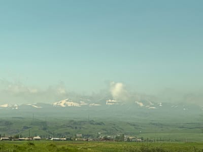 The image depicts a panoramic view of a rural landscape near Sevan, Armenia, with Mount Ararat faintly visible in the distance. In the foreground, a green field stretches out, with scattered houses and farm structures of varying sizes and colors dotting the middle ground. Several utility poles are visible throughout the landscape. The background is dominated by rolling green hills leading to a majestic mountain range partially covered in snow and shrouded in mist. The sky is a clear, light blue with soft, diffused clouds. The overall impression is one of serene natural beauty and rural tranquility.