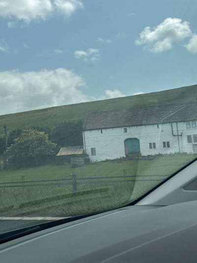 The image is taken from a car driving down a rural road, with a view of a single-story white building with a blue door. The building has a brown roof and windows on the side and is located on a grassy hillside. There is a fence along the edge of the road. The sky is clear blue with scattered white clouds. The image was taken near the village of Wadsworth, Calderdale, United Kingdom. The coordinates (53.643, -1.948) correspond to a location near the village of Wadsworth, Calderdale, United Kingdom.