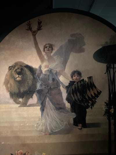 The image shows a large, semi-circular mural depicting a scene with allegorical figures and a lion. In the center, a woman with fair skin, light brown hair styled in loose waves, and a light complexion stands with her right arm raised, holding a laurel wreath. She wears a flowing, light blue and white gown. To her left, a large, powerful lion with a mane of tawny fur stands facing forward, looking directly at the viewer. To the woman's right, a young child with curly brown hair and a fair complexion, dressed in dark clothing, holds a shield with a diamond pattern of black and gold, and a laurel wreath. The figures are positioned on a set of stone steps. In the background, a faint shadow of a hand or claw is visible, and to the right, a dark, elaborate metal structure, possibly a candelabra or sconce, with floral arrangements. The mural is mounted within a dark frame, and part of a dark ceiling is visible at the top. This mural is located in the Hall of Sessions of the Federal Palace in Bern, Switzerland.