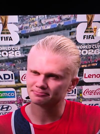 The image is a close-up, slightly angled shot of a young man with blond, slicked-back hair, wearing a red shirt with blue and white trim. He has a fair complexion and is looking slightly to his left with a neutral expression, his lips slightly parted. In the background, there are large digital billboards displaying various logos and text, including "FIFA WORLD CUP 2026," a gold trophy image, "Lenovo," and "Coca-Cola." The overall environment appears to be a large-scale event or stadium setting, suggested by the presence of multiple advertisements and a blurred crowd in the distance. The provided location of Kosshy, Kazakhstan, is not verifiable from the visual information within the image itself.