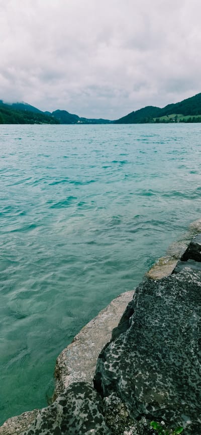A serene, wide-angle shot captures a body of water with rolling green hills and a cloudy sky in the background. The foreground features a rocky shoreline made of large, dark, and light gray stones. The water is a vibrant turquoise, with small ripples on its surface, indicating a calm day. The scene is devoid of any people or specific landmarks that would definitively place it in Brunn, Austria, or any other identifiable city. The overall impression is one of natural beauty and tranquility.