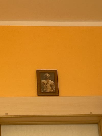 A framed picture hangs on an orange wall next to a doorway. The picture appears to be a painting or drawing depicting two figures, one of whom is cradling a smaller figure. The frame is dark brown and rectangular. The doorway is to the right of the picture and is framed by a lighter colored trim. A closed door with a translucent panel is partially visible within the doorway. The lighting suggests it is daytime, as natural light seems to be coming from the right side of the image. The overall setting appears to be indoors, possibly a hallway or room in a home. There is no visible text in the image.