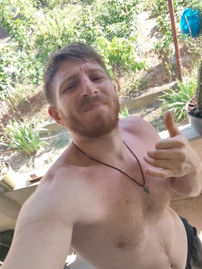 The image is a selfie of a shirtless man with reddish-brown hair and beard, giving a thumbs-up. He is wearing a dark necklace with a cross pendant. The man appears to be outdoors, with lush green foliage and a wire fence visible in the background. A large blue barrel and a cactus plant are also present. The man is standing on a concrete surface. The image appears to have been taken in a backyard or garden setting. Given the context provided, the location is identified as Florești, Moldova.