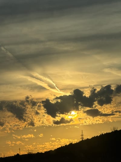This is a vertical, landscape photograph of a dramatic sunset over Kaspichan, Bulgaria. The sun, a bright orb tinged with yellow and orange, is partially obscured by dark, billowing clouds. The sky is a canvas of warm hues, transitioning from a deep golden yellow near the sun to lighter, almost gray tones on the left side of the frame. Wispy, elongated contrails streak across the sky, adding a dynamic element to the scene.

In the foreground on the right, the silhouette of a dark, tree-lined hill or embankment runs diagonally from top to bottom. Two electricity pylons are visible against the bright sky, indicating the presence of power lines and infrastructure. The overall mood is one of awe and a touch of melancholy, characteristic of a powerful sunset. The time of day is clearly sunset, given the low angle of the sun and the warm, diffused light. The weather appears to be fair, with clouds contributing to the dramatic visual spectacle. No text is visible in the image.