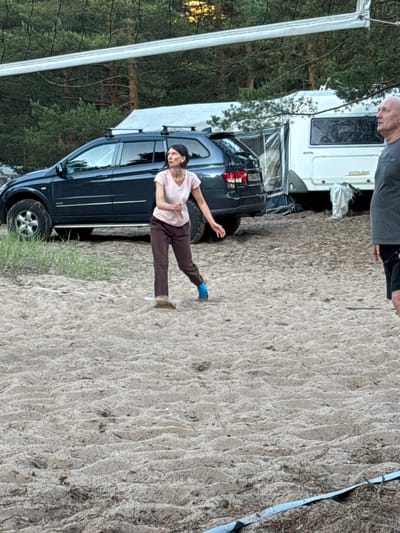 A woman in a light pink t-shirt and brown pants is captured mid-action in a sandy area, possibly playing volleyball, with a dark SUV and a white camper trailer visible in the background, amidst a backdrop of pine trees. Another man, wearing a grey t-shirt and black shorts, stands to the right, looking away from the camera. The ground is sandy and uneven, with a volleyball net strung across the top of the frame. The scene appears to be a campsite, potentially located in or near Ozerki (Krasnodolinskaya), Russia, given the presence of the camper and the natural environment.