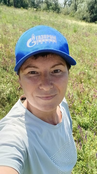 A woman wearing a blue baseball cap with the "Gazprom" logo and a light blue t-shirt takes a selfie outdoors in a grassy field with trees in the background. The woman has short brown hair and is smiling. The image appears to have been taken in Nadezhdenka, Russia.