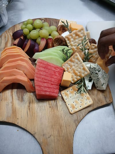 A charcuterie board is displayed. The board features various cheeses (including a block of blue cheese), crackers, grapes, cut plums, sliced honeydew melon, a chunk of watermelon, and slices of cantaloupe. A person's hand is reaching towards the cheese on the upper left of the board. The board itself is made of wood. The setting is unknown. There is no information provided that allows me to describe the location in Nwanaeanoka, Botswana.