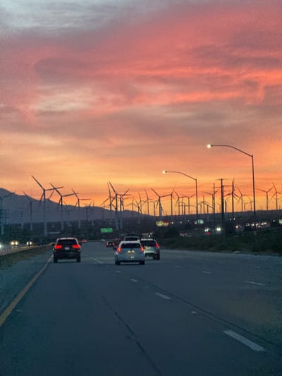 The image captures a vibrant sunset over a highway lined with a vast wind turbine farm. The scene is set in what appears to be a desert or semi-arid landscape, consistent with the location context of Desert Hot Springs, United States, where wind farms are prevalent.
The sky is the most striking feature, painted with brilliant hues of fiery orange and red closest to the horizon, gradually transitioning to soft pinks and lighter grays higher up, with hints of blue at the very top. These colors clearly indicate the time of day is sunset or twilight.
In the middle ground and background, numerous wind turbines stand silhouetted against the colorful sky. Their tall structures and rotating blades stretch across the landscape, extending into the distance on both sides of the highway. Mountains are faintly visible as dark shapes behind the wind farm.
In the foreground, a multi-lane highway stretches forward, appearing dark gray under the low light. Several cars are visible on the road, with their taillights glowing red. The perspective suggests the photo was taken from inside a moving vehicle, looking ahead. The vehicles include sedans and SUVs. Streetlights with bright white lamps are illuminated along the highway, casting pools of light.
On the left side of the highway, in the middle distance, a green road sign is visible, but the text on it is not legible. The overall impression is one of modern infrastructure coexisting with natural beauty, highlighting renewable energy production during a beautiful natural phenomenon. No specific individuals or faces are discernible.