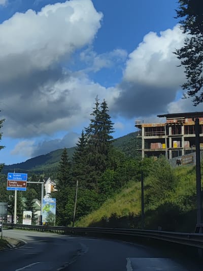 This media file depicts a mountainous landscape in Borșa, Romania, on a partly cloudy day. In the foreground, a wet asphalt road curves to the right, bordered by a metal guardrail. To the left of the road, a blue directional sign points left towards "Iacobeni" and "Cărlibaba" and then right towards "Complex Turistic Borșa." Next to this sign, a large billboard displays a person in a bright green ski suit, with the text "THE SEASON STARTS HERE" at the top.

Further to the left, several houses with pitched roofs are visible nestled amongst trees. A second, smaller billboard with black and white imagery is also present. The middle ground is dominated by a steep, grassy hill dotted with trees. Atop this hill, a multi-story building is under construction, with visible concrete floors, scaffolding, and some yellow signage. The structure appears to be a large hotel or resort.

In the background, lush green mountains rise, partially obscured by dramatic, dark grey and white clouds against a bright blue sky. Tall evergreen trees are scattered throughout the scene, particularly on the left and right sides. The overall impression is one of a developing tourist area in a natural, scenic setting. The construction of the building suggests ongoing development for tourism, likely related to the nearby ski resort, as indicated by the billboard. The weather appears to be dynamic, with both sunshine and heavy clouds present.