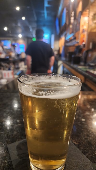 A close-up, slightly high-angle, and horizontally oriented photograph depicts a glass of beer with a frothy head, placed on a dark, textured countertop. The background suggests a bar setting.
Foreground: The beer glass is the primary focus, showcasing a clear, golden liquid topped with white foam. The glass is positioned slightly off-center, allowing a view of the beer's carbonation. A dark-colored cloth or napkin is partially visible beneath the glass.
Background: The background features a blurred bar scene, indicated by bar shelves, a bartender, and several patrons. Lighting elements, like illuminated signage or decorative lights, create bright spots within the blurred context.
The location in Jackson, United States of America, is not discernible from the visual elements.