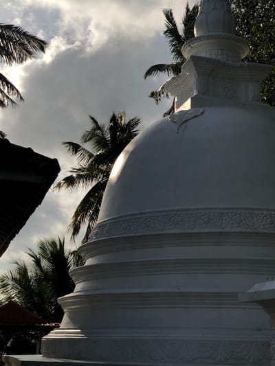 This image shows a white stupa, a Buddhist reliquary, in Pilana, Sri Lanka. The stupa is partially visible, with its dome and tiered base dominating the foreground. It is decorated with intricate carvings, including floral motifs. Palm trees frame the stupa, and a cloudy sky with patches of sunlight creates a dramatic backdrop. To the left of the stupa, the dark, tiled roof of a building is visible, suggesting its proximity to a temple or other religious structure. The scene is bathed in soft, natural light, likely from the late afternoon sun.