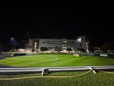 Captured at night, the image presents a view of Kempton Park Racecourse in the Borough of Spelthorne, United Kingdom. The primary focus is a large green expanse, possibly a portion of the racecourse infield or surrounding area, illuminated by artificial lighting. A sprinkler system is actively watering the grass. A yellow caution tape is stretched across the green. A building, likely part of the racecourse facilities, stands in the background. A hose and other equipment are visible in the foreground, and a small crowd is visible near the building. The overall scene suggests preparations or maintenance activities at the racecourse during the evening.