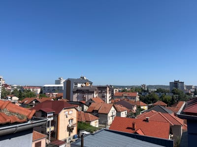 The image captures a panoramic view of a residential area in the city of Cacak, Serbia, under a clear blue sky. The foreground features a section of a building's roof with weathered orange tiles and a grey gutter. Beyond this, a dense collection of houses with predominantly red tiled roofs fills the mid-ground. These houses vary in architectural style and size, with some featuring balconies, satellite dishes, and air conditioning units. Several multi-story buildings, including apartment complexes and what appears to be an office building, are visible in the background, interspersed with trees and other structures. The overall impression is of an urban or suburban landscape on a sunny day. There are no people or vehicles visible in the image.