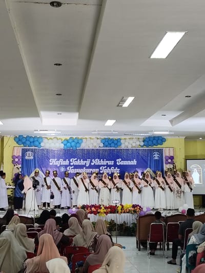 The image depicts an indoor event, likely a graduation or awards ceremony, taking place in a venue in Kutalhoksukon, Indonesia. A group of approximately 18 young people, dressed in white robes and some wearing golden crowns, are standing on a raised stage. They appear to be the honorees. Behind them, a large blue banner with white text reads "Haflah Takhrij Akhirusannah & Tasyaquran Tahfidz," indicating the nature of the event. The stage is decorated with balloons and floral arrangements. In the foreground, an audience is seated, many of them wearing headscarves, facing the stage. The lighting in the room appears to be from overhead fluorescent panels and spotlights. The overall atmosphere suggests a formal and celebratory occasion within a school or community hall setting.