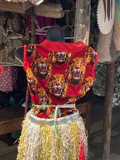 In an outdoor market stall in Afikpo, Nigeria, a headless torso mannequin is positioned centrally, facing away from the viewer. The mannequin displays a sleeveless red top featuring multiple repeating patterns of roaring lion heads and a red collar. Around the mannequin's waist, a skirt-like garment is tied with a green string; this garment is composed of dense, shredded strips of material, predominantly yellow and white, creating a fringed texture. Above the mannequin, a rough wooden beam of a stall structure is visible. In the background, various patterned fabrics are displayed, including dark fabric with light circular motifs and pink fabric. A weathered vertical wooden pole and a partially visible, irregularly shaped white object with a dark border are present on the right. A wooden bench or table is partially visible on the left. The scene is illuminated by daylight.