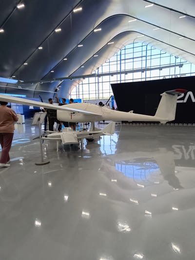 In a large, modern exhibition hall with a curved, dark ceiling illuminated by rows of white lights, a light-colored drone is displayed on a polished, reflective gray floor. The drone has a long, slender fuselage and a prominent horizontal stabilizer at the rear. It is supported by landing gear, and what appears to be a second, smaller drone or a component of a drone is situated beneath it. Several people are standing around the drone, some observing it and others engaged in conversation. One person in the foreground is partially visible, wearing brown attire and white shoes. In the background, a large black banner displays a white logo that appears to be the letters "ZALA," possibly indicating the manufacturer or a sponsor. The rear wall of the hall features large, grid-like windows that allow natural light to enter and reflect on the floor. The scene suggests an aerospace or technology exhibition. This image was taken in Korsakov, Russia.