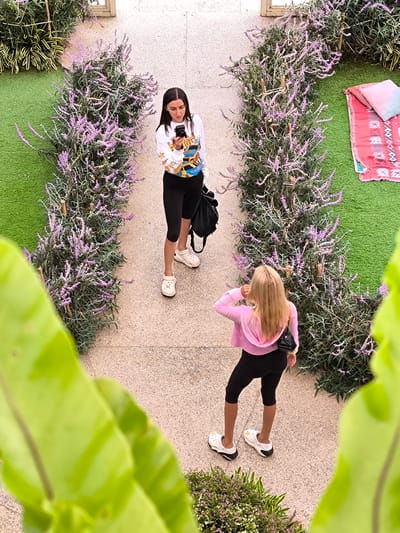 The image, taken from an elevated perspective, shows two women standing on a stone-paved path bordered by flowering plants with purple blossoms. The location is likely Candikuning, Indonesia based on the flora and possible Balinese-style architecture present.

One woman, with dark hair, wears a long-sleeved white top, black cycling shorts, and white clogs. She is holding and looking at a camera. She also has a black shoulder bag.

The other woman, with long blonde hair, is wearing a light pink hoodie, black cycling shorts, white clogs with black socks, and a small black purse over her shoulder. She is facing away from the camera and appears to be adjusting her collar or hair.

On one side of the path, a section of green turf is visible with a patterned rug and pillows on it. The image appears to be taken outdoors during daylight. A large green plant with large leaves is partially visible in the foreground of the image, obstructing the view. The flowering plants on either side of the path appear to be arranged to create a sort of archway. The background is slightly blurred, but appears to be a wall or structure.