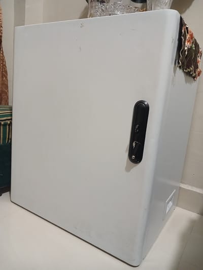A large, white, chest freezer is positioned in the corner of a room. The freezer has a black, elongated handle with a keyhole and a lever mechanism. To the left of the freezer, a green and dark striped cushion with a gold trim is visible. A section of a brown patterned curtain is also present on the far left. On top of the freezer, a decorative floral cloth is draped over the right edge. The floor appears to be tiled in a light color. In the background, a wall with vertical lines is visible. A white rectangular vent is present on the right side of the freezer near the floor. The image does not depict any individuals or actions. The specific location of Geudonggeudong, Indonesia, is not indicated by any visual elements in the image.