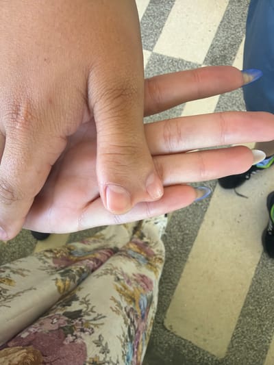 The image shows a close-up view of two hands intertwined. One hand, on the left, is larger and appears to be resting palm-up. The other hand, on the right, is placed over the first, with its fingers extending upwards. The fingers of the second hand have long, pointed, light blue acrylic nails. The background consists of a tiled floor with a pattern of alternating light and dark grey squares, and a glimpse of patterned fabric on what appears to be a person's lap. The overall impression is of two hands in a gentle clasp. There is no identifiable information about the city of Dinkata, Bulgaria, in the image itself.