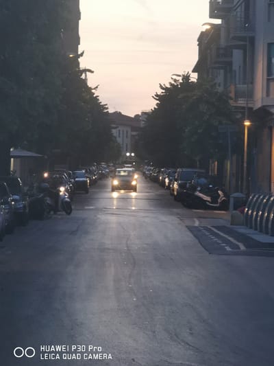 The image depicts a street scene in Follonica, Italy, at dusk. A street is lined with parked cars and motorcycles on the left and buildings on the right. The sky is a pale orange and pink, indicating the sun has recently set. A yellow car is driving down the street, its headlights illuminated. The street is paved and appears to be wet, reflecting the lights of the car and street lamps. The overall mood is peaceful and quiet, suggesting a typical evening in a small Italian city.