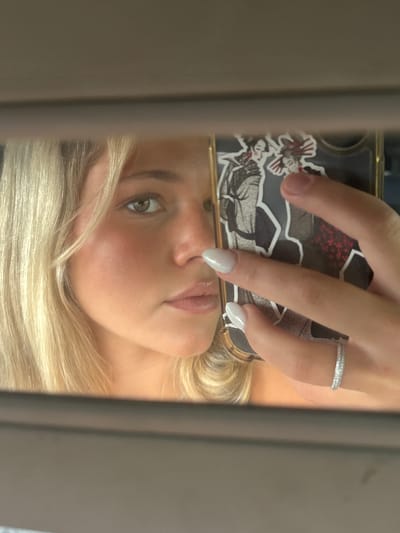 A person is reflected in a mirror while holding a smartphone. The smartphone has a decorated case with multiple stickers, and a finger wearing a ring is visible. The person has blond hair and green eyes. The location is not discernible from the image.