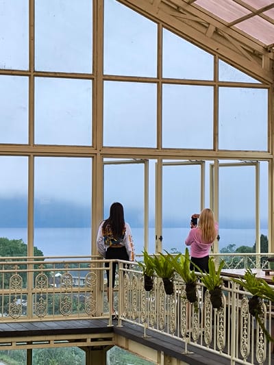 Two women are standing on a bridge, looking out over a lake. The woman on the left has long dark hair and is wearing a white shirt with a colorful design and black leggings. The woman on the right has long blonde hair and is wearing a pink hoodie and black leggings. The women are in an enclosure with large glass windows. The window panes are open to let in fresh air. Outside, the lake is surrounded by lush greenery. The bridge has an ornate railing with hanging ferns. The scene is in Candikuning, Indonesia.