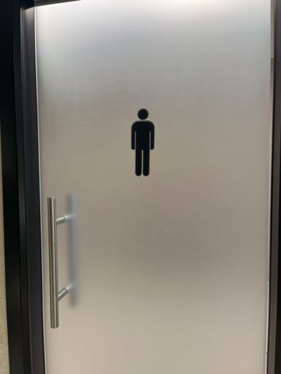 The image shows a frosted glass door, likely for a restroom, located in Dorno, Italy. A prominent black, stylized male figure icon is adhered to the upper-left to central portion of the frosted glass. This pictogram, which appears to be lying on its left side, features a circular head and a rectangular body with two parallel lines forming the legs, commonly used to designate a men's facility.
The door is framed in black material, and a brushed metal, horizontal bar-style handle is visible on the lower right side, attached by two vertical supports. The frosted nature of the glass provides privacy, obscuring the view behind the door while still allowing light to pass through. The lighting suggests an indoor setting, likely during the day or under artificial light. There is no visible activity taking place, and no additional text or specific location cues are present beyond the door and its handle. The overall design implies a functional and modern aesthetic, typically found in public or commercial buildings.
