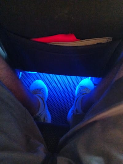An overhead view shows a person's feet wearing white sneakers, positioned inside what appears to be a vehicle's footwell. The footwell is illuminated by blue LED lights, casting a vibrant glow on the floor, which has a textured, perforated surface. A red item, possibly a piece of clothing or a bag, is visible tucked into a compartment above and behind the feet. The surrounding interior of the vehicle is dark and out of focus. The scene is static and appears to be a casual snapshot.