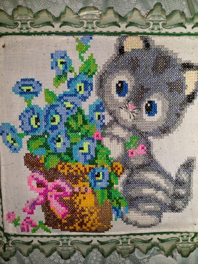This is a close-up, eye-level photograph of a cross-stitched embroidery of a gray tabby kitten sitting next to a potted plant with blue flowers. The kitten has large blue eyes, pink accents on its nose and ears, and is gray with darker gray stripes. The plant is in a brown pot with a pink bow and some smaller pink flowers at its base. The embroidery is framed by a decorative green border. The image is brightly lit and in focus, highlighting the detailed stitching. There is no indication of location within the city of Shpychyntsi, Ukraine.