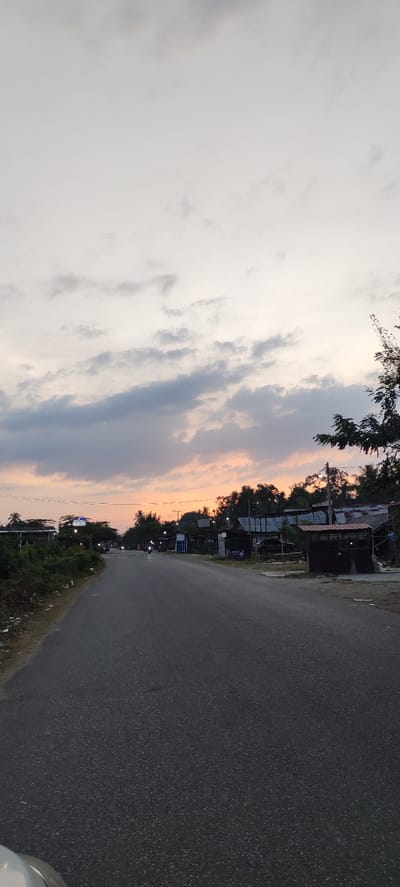 This is a vertical, eye-level, medium shot captured at dusk on a road in Sumbokrayek, Indonesia. The sky is a pale grey with scattered clouds tinged with orange and pink from the setting sun, creating a soft glow. A dark asphalt road dominates the lower half of the image, stretching into the distance and appearing wet. On the left side of the road, lush green vegetation borders the asphalt, and on the right, a line of buildings with corrugated metal roofs and some wooden structures are visible, suggesting a village or small town. A few vehicles, including motorcycles and a car, are present on the road, and some streetlights are illuminated. The overall atmosphere is peaceful and serene, typical of a rural Indonesian village at the end of the day.