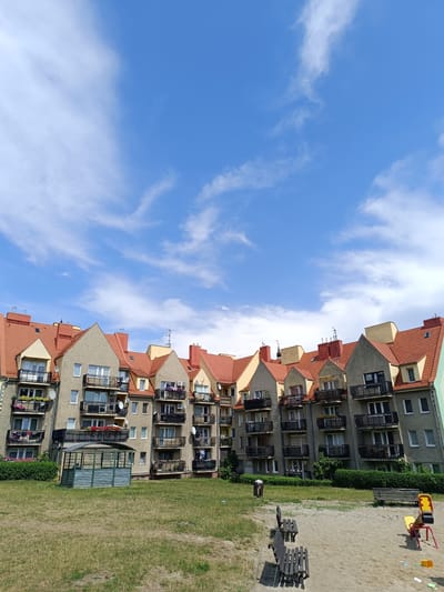 The image shows a row of multi-story apartment buildings with red tiled roofs against a bright blue sky with scattered white clouds. The buildings have a light beige facade, with balconies on each level. There is a small, green, open-sided shelter with a metal roof in front of the buildings, situated on a grassy area. In the foreground, there is a sandpit with several wooden benches and a small playground structure with yellow and red components. The scene appears to be a residential area in the city of Dzierżoniów, Poland. No people are visible in the image.