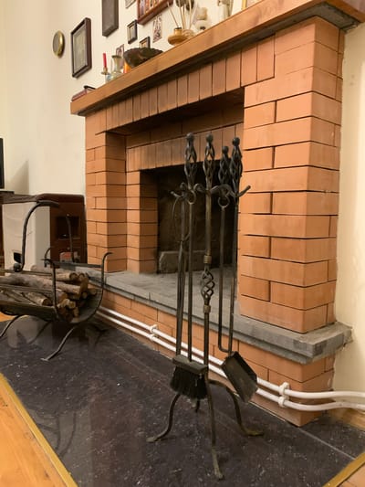 The image shows a brick fireplace with a wooden mantelpiece. On the mantelpiece, there are various decorative items, including framed pictures, a candle, and a vase. In front of the fireplace, there is a set of fireplace tools including a poker, tongs, a brush, and a shovel, arranged on a metal stand. To the left of the fireplace, a metal log holder is filled with firewood. There is also a white and brown heating appliance next to the log holder. The floor in front of the fireplace is a dark, speckled material, and a wooden floor is visible to the left. White pipes are mounted on the wall behind the fireplace tools. The overall setting appears to be an indoor living space. The location of Mepiskalaki, Georgia, is not directly evident from the image itself.