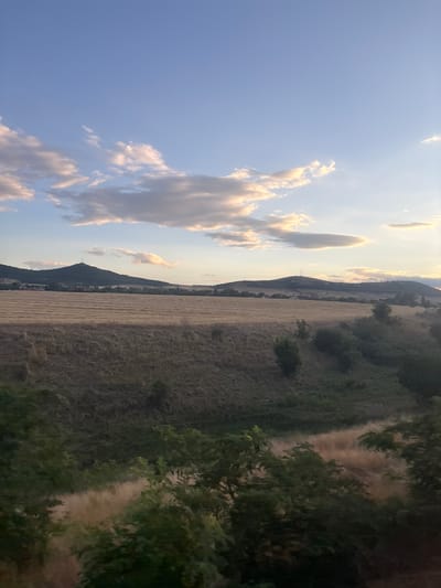 The image depicts a rural landscape under a partly cloudy sky, possibly taken during sunset or sunrise due to the warm light illuminating the clouds. The scene features rolling hills with a large field of harvested crops in the mid-ground. A dark, conical hill with a tower on its summit is visible in the distance, silhouetted against the sky. The foreground is dominated by green foliage, suggesting trees and bushes, some of which are blurred, indicating movement during the capture of the image. The overall impression is one of a tranquil, natural environment, possibly located outside a populated area.