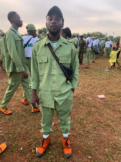 The image depicts an outdoor scene in an open, reddish-brown field in Kubi A, Nigeria, under a hazy, overcast sky. The foreground is dominated by a dark-skinned adult male standing centrally, facing the camera. He wears a light green National Youth Service Corps (NYSC) uniform consisting of a long-sleeved shirt with rolled cuffs and matching trousers, paired with bright orange ankle boots, black laces, black soles, and white socks. A dark green baseball cap is worn backward, and a black sling bag with a red and black strap is worn across his chest. He sports a short beard and a white bracelet on his right wrist.
In the midground and background, numerous other individuals are visible, predominantly young adults also dressed in similar light green NYSC uniforms. Many wear white t-shirts with "NYSC" printed in green on the back, and some have matching green caps. They are arranged in rows or loose formations, suggesting an organized event. One individual is seen carrying a black trash bag, and a person in a camouflage uniform stands among the NYSC members. On the far right, several children are present, including one wearing a bright yellow garment. The ground is sparsely covered with dry grass and exhibits scattered debris, including plastic bottles. Distant trees and faint structures are visible at the horizon line.