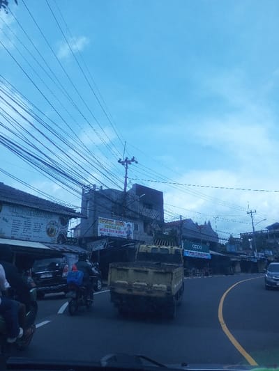This image depicts a bustling street scene in the city of Chori, Nigeria, under a bright blue sky with scattered white clouds. A dense network of black utility wires spans across the upper portion of the frame. The foreground shows an asphalt road curving to the right, marked by a yellow dividing line.
Vehicles are actively navigating the street. In the left foreground, the rear of a dark-colored SUV or minivan is partially visible, with a person on a black motorcycle, wearing a dark jacket and helmet, carrying a blue package on the rear, positioned behind it. Further right, a yellow, open-back dump truck with a visible rear license plate is driving away from the viewer. Another dark-colored vehicle is visible further down the road on the right curve.
Along both sides of the street, commercial buildings line the thoroughfare. On the left, a building features a large white sign with text in the Indonesian language, including "WARUNG PEAYEM CAKRA." Below it, an awning extends over a visible parked vehicle. Adjacent to this, another building displays a sign with Indonesian text such as "RENTAL MOBIL DAN MOTOR" (Car and Motorcycle Rental) and "MODERN WARGA." Utility poles with multiple cross arms support the overhead wiring at various points along the street. On the right side of the street, a building prominently displays a blue sign with the brand name "OPPO" in white lettering. The architectural styles of the visible structures suggest an urban commercial environment, with details consistent with Southeast Asian design elements. The overall scene indicates active daily commerce and traffic.