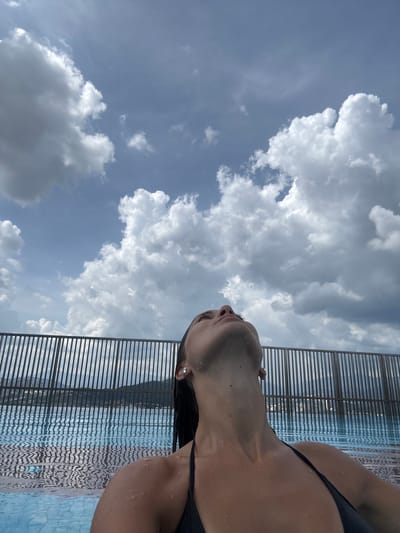 The image captures a close-up, low-angle view of a woman relaxing in a swimming pool, oriented vertically with the sky dominating the left side. The woman's upper torso, neck, and face, from below the nose, are visible on the right side of the frame. She wears a dark swimsuit, and her skin is visibly wet with water droplets. Her head is tilted back, with her gaze directed upwards towards the sky.
The setting is an outdoor swimming pool, with the water's surface, showing ripples and reflections, visible alongside the woman. A modern, dark-colored slatted fence or railing runs horizontally across the top-right portion of the image, bordering the pool area. The background is a bright blue sky filled with numerous large, fluffy white and grey cumulus clouds, indicating a partly cloudy day. The bright lighting suggests it is daytime. The overall impression is one of relaxation and contemplation. No text is visible in the image. The location is Ampang Jaya Municipal Council, Malaysia.