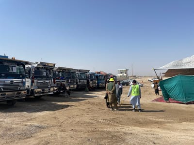 The scene depicts an outdoor, unpaved area in Pir Dust, Afghanistan, under a clear blue sky. A prominent line of at least eight heavy-duty dump trucks, primarily blue and white/silver, is parked on the left side of the frame, facing left. The trucks feature chrome detailing and some have Arabic script on their sun visors. In the midground, a group of approximately five men are standing on the dry, reddish-brown dirt. One man in the foreground wears a light blue traditional outfit with a bright yellow-green high-visibility vest and appears to be interacting with another man wearing a yellow-green wide-brimmed hat and olive-green traditional attire. A long, yellow measuring tape or similar object is visible near their interaction. In the distant background, a small, light-colored building with a blue roof stands on a barren horizon. To the right, several passenger cars are parked, and a large, light-colored net structure provides shade over an indistinct mound. Closer to the foreground on the right, a dark green canvas tent is erected on red ground coverings, secured with ropes. The ground is consistently dry, dusty, and marked with tire tracks.