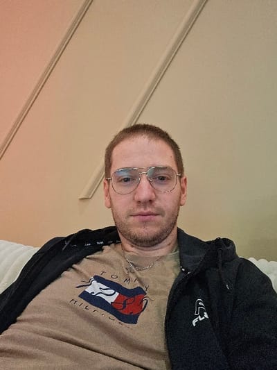 An adult male is depicted in a frontal, chest-up shot. He has short light brown hair and light facial stubble, and wears silver-framed aviator-style glasses. He is dressed in a tan t-shirt featuring a red, white, and blue "Tommy Hilfiger" logo, a silver-colored chain necklace with a small pendant, and a partially unzipped dark-colored zip-up hoodie with a white "Fila" logo visible on the right chest area. He appears to be seated on a light-colored, textured surface, possibly a sofa. The background consists of a plain, warm peach-toned wall with two parallel decorative moldings running diagonally upwards on the left side. The scene is located indoors in the city of Dragushinovo, Bulgaria.