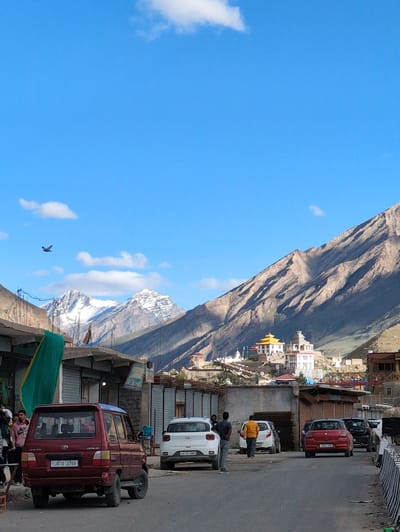 In Padum, India, a scene unfolds under a bright blue sky dotted with scattered white clouds. A dirt road runs through the foreground, lined with small buildings and shops. Several cars are parked along the road, including a red SUV in the left foreground and a white hatchback to its right. A person in a pink hoodie stands near the red SUV, looking away from the camera. Further down the road, two men are walking away from the camera. In the background, a cluster of white and yellow buildings, possibly a monastery, is nestled on a mountainside. The imposing, rocky slopes of the Himalayas dominate the upper right portion of the image, with snow-capped peaks visible in the distance. A bird is captured in mid-flight against the vast expanse of the sky.