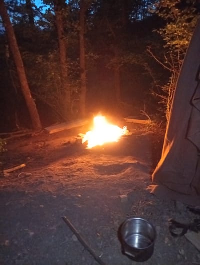 A night-time scene depicting a campsite with a campfire in what appears to be a wooded area. The fire is brightly lit, illuminating the surrounding ground and nearby trees. A dark, tent-like structure is visible to the right of the frame. In the foreground, there is a metal cooking pot on the ground. A stick is also lying on the ground near the pot. The location is specified as Malko Chochoveni, Bulgaria.