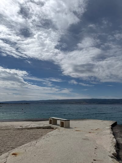 A concrete bench sits alone on a concrete platform overlooking the Adriatic Sea in Dugi Rat, Croatia. The bench is situated near the edge of the platform, which slopes gently down to the water. The sea is a deep blue, with light ripples on the surface. The sky is bright blue with scattered white clouds. The horizon is defined by a line of hills in the distance. The scene is devoid of people.