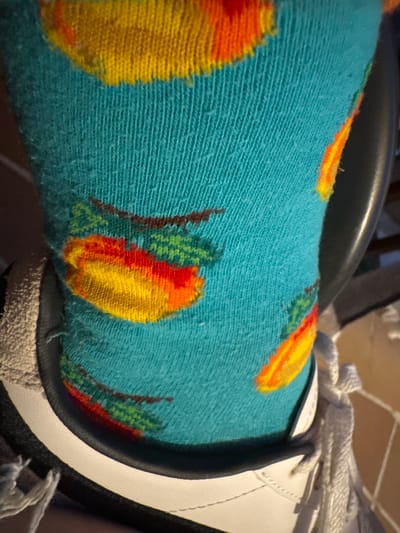 The image is a close-up, eye-level shot of a person wearing a teal-colored sock with a pattern of peaches. The sock is knitted and appears slightly fuzzy. Parts of a white sneaker are visible at the bottom of the frame, with white laces. The background is dark and out of focus, suggesting an indoor setting. The image does not provide any visual information that would allow for a determination of its location within El Escorial, Spain.