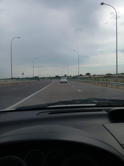 The image depicts a view from inside a vehicle looking forward onto a multi-lane highway. The sky is overcast with scattered clouds. Streetlights line both sides of the highway at regular intervals. Several vehicles are visible on the road, including a white sedan in the distance on the right lane and another white car in the left lane ahead. The highway appears to be elevated or on a causeway. On the right side of the highway, there is a metal guardrail, and beyond that, a fence and some buildings and trees are visible. On the left side of the highway, there is also a guardrail, and further left, what appears to be a body of water or flat land with vegetation. The foreground shows the interior of the car, including the dashboard and windshield. The scene is consistent with a highway in Eskikurgan, Uzbekistan, given the architectural style of the buildings and the general landscape.