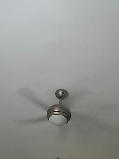 The image depicts an upward view of a modern ceiling fan with an integrated light fixture, centrally positioned against a plain white ceiling. The fan's main body and downrod are constructed from a brushed metallic material, likely brushed nickel or silver-toned. A translucent white, spherical or dome-shaped light cover is affixed to the bottom of the fan's motor housing. The fan blades are in motion, evidenced by their blurred appearance and the diffused, sweeping shadows they cast on the white ceiling above and to the left of the fan's body. No other objects or individuals are visible. The scene is located within a structure in Πλατύς Γιαλός, Greece.