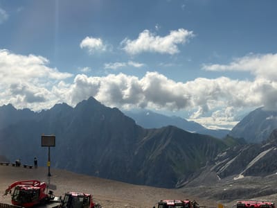 This image captures a high-altitude mountain landscape, likely in the Austrian Alps near Wildermieming, given the presence of rugged, snow-capped peaks and a vast, rocky terrain. The sky is a bright blue, dotted with scattered white clouds, suggesting fair weather. In the foreground, two bright red snow groomers or similar specialized vehicles are parked on a gravelly surface. One is closer to the viewer, and the other is partially visible to its right. Further back, a single lamppost stands with a rectangular sign and two orange lights on top. Two people are visible near the lamppost: one is seated on a bench, and the other is standing. The background is dominated by a dramatic mountain range with steep slopes and visible rock formations, some patches of snow are also present on the mountainsides. The overall scene conveys a sense of remote, mountainous wilderness, possibly a viewpoint or operational area for mountain transport or maintenance.