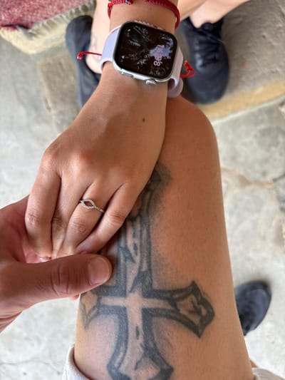 The image shows a close-up view of two people holding hands. One person is wearing a white smartwatch on their left wrist, and a diamond ring on their left ring finger. The other person's hand is visible reaching in from the bottom left of the frame. The background consists of blurred legs and feet, with one person wearing black sneakers and the other wearing dark shoes. A large cross tattoo is visible on the leg of the person wearing the smartwatch. The scene appears to be outdoors, possibly in a public space, given the presence of paved ground and casual footwear. The image is shot from a slightly elevated angle, focusing on the hands and arms.