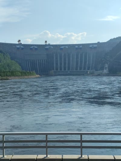 The image depicts a wide, blue river flowing in the foreground, with a large, imposing concrete dam stretching across the background. The dam is a colossal structure, characterized by numerous vertical concrete pillars or spillways, and topped with a flat deck. It is adorned with large circular emblems featuring the colors of the Russian flag: white, blue, and red. Two prominent red cranes are visible on the upper edge of the dam.
To the left of the dam, a lush green forested hillside slopes down towards the water. On the right side of the dam, there are power lines and a tall, lattice-like transmission tower. The water in the river is rippled, suggesting a gentle current.
In the foreground, a metal railing with three horizontal bars runs across the bottom of the frame, supported by vertical posts. This railing, along with concrete blocks beneath it, suggests a viewing platform or promenade overlooking the river and the dam.
The sky above is a pale blue with scattered white, fluffy clouds, indicating fair weather. The lighting suggests it is daytime, likely midday or afternoon, given the brightness and lack of long shadows. The overall impression is one of immense scale and man-made power, set against a natural landscape. The presence of Russian flags on the dam points to its location in Russia, specifically Cheremushki, Russia, as per the provided location context. No visible text is present in the image.