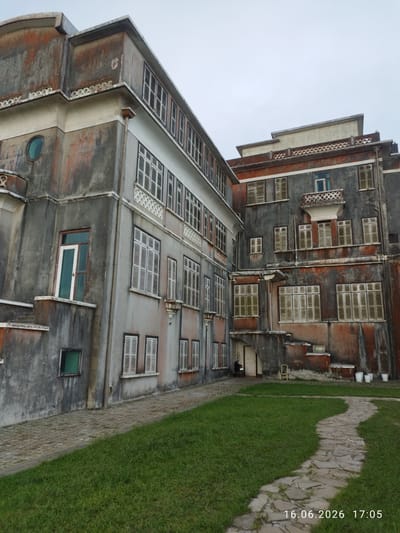 The image depicts an exterior view of a weathered, multi-story building in Phumi Pôpôk Vil, Cambodia. The architecture features a combination of grey and reddish-brown concrete, with numerous windows, many of which are covered with shutters. Some sections of the building show signs of aging, including peeling paint and staining. A paved pathway winds through a grassy area in front of the structure. In the background, there are a few white buckets and a motorcycle parked near an entrance. A date and time stamp, "16.06.2026 17:05", is visible in the lower right corner.