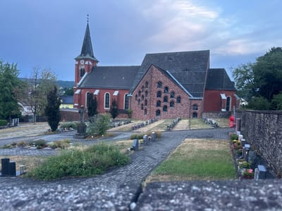 The image depicts a cemetery adjacent to a red brick church in Wadern, Germany. The sky is overcast with a pale blue hue, suggesting either early morning or late evening. The church features a tall, pointed steeple with a cross at its apex and a clock face. The main body of the church is constructed of red brick, with arched windows visible. An additional section of the church is built with reddish-brown brick and features a repeating pattern of arched openings. The cemetery is laid out with rows of gravestones and small floral arrangements. Paved pathways wind through the cemetery. A stone cross stands prominently in the foreground of the cemetery. Greenery, including trees and bushes, is present throughout the scene. A stone wall borders the right side of the cemetery. No people are visible in the image.