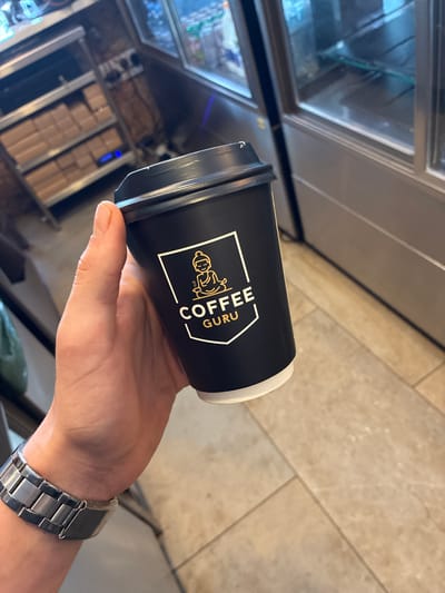 A person's hand is holding a black coffee cup with a white and gold logo. The logo depicts a meditating figure with the words "COFFEE GURU" written below. The cup has a black lid. The person is wearing a silver metal watch on their wrist. The background shows a tiled floor and what appears to be shelves with boxes. The scene is indoors, likely a cafe or shop. The image does not contain enough information to determine its location in Kato Polemidia Municipality, Cyprus.